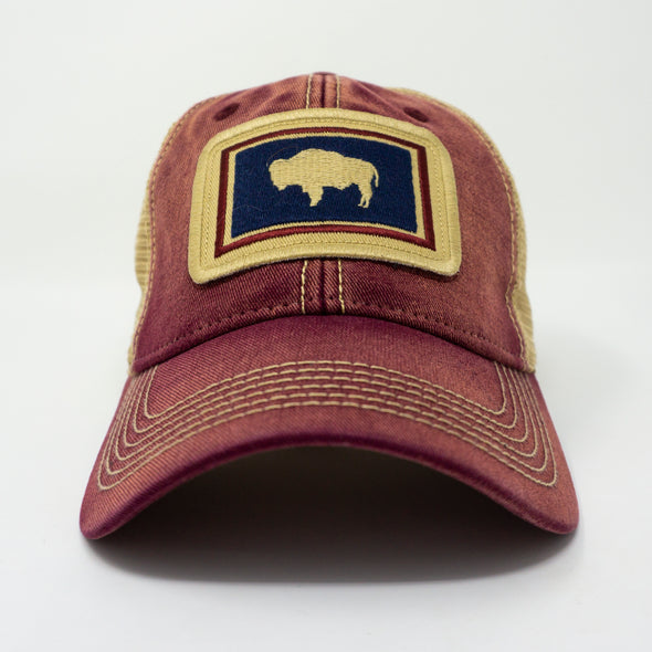 Brick red colored trucker hat with khaki mesh panels on the back and tan stitching. Ballcap is embroidered with the Wyoming flag. The patch depicts a natural Bison inside of a dark blue rectangle with a red border.