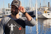 Man with beard wearing a black trucker hat with embroidered Blackbeard pirate flag patch on the center and khaki mesh back panels at marina. Patch depicts a natural colored skeleton with horns piercing a red bleeding heart with a spear. 