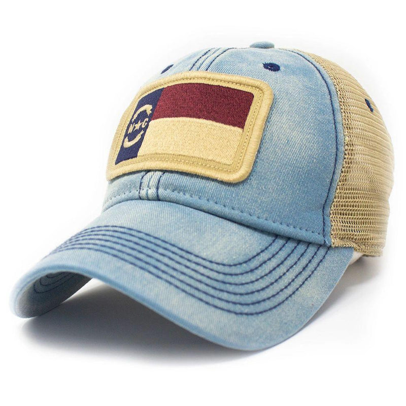Trucker hat with khaki colored mesh backing, light denim cotton front panel and bill with tan stitching. Ballcap has an embroidered patch of the North Carolina flag in in the center.