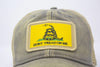 Close up of grey trucker hat with khaki mesh back panels and tan stitching laying. Ballcap is embroidered with a patch of the Gadsden flag, the patch has a yellow background with a snake coiled in grass and the words Don't Tread On Me
