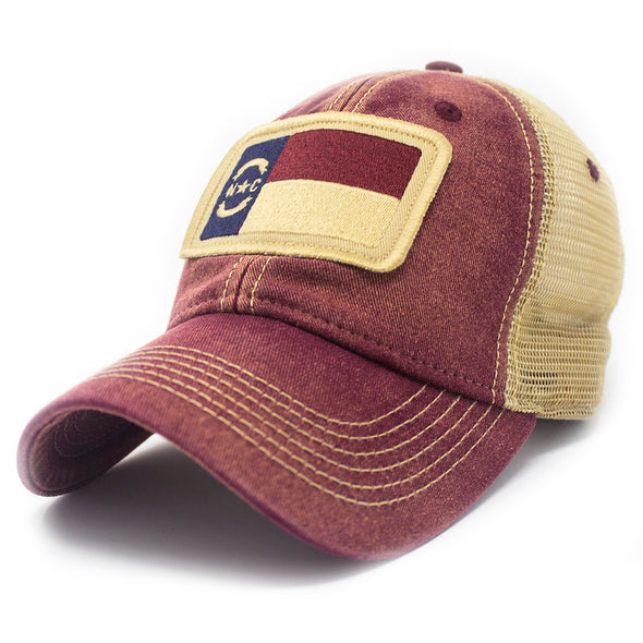 Trucker hat with khaki colored mesh backing, dark red cotton front panel and bill with tan stitching. Ballcap has an embroidered patch of the North Carolina flag in in the center.