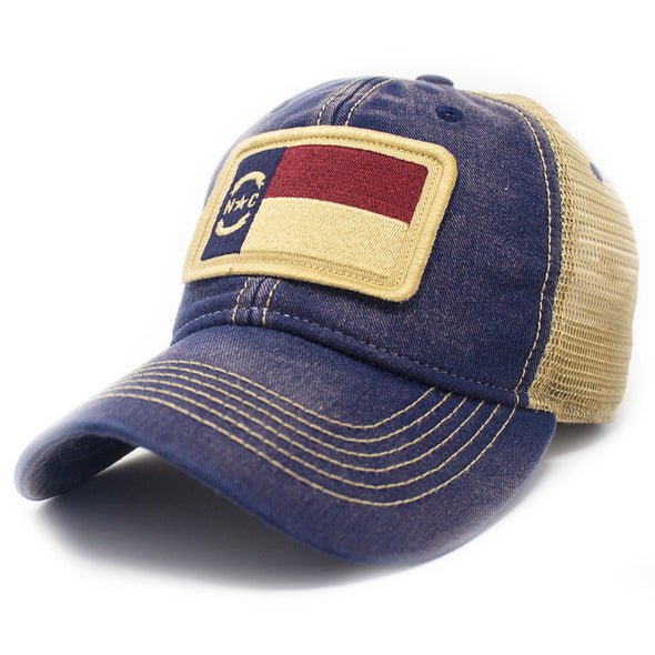 Trucker hat with khaki colored mesh backing, dark denim cotton front panel and bill with tan stitching. Ballcap has an embroidered patch of the North Carolina flag in in the center.